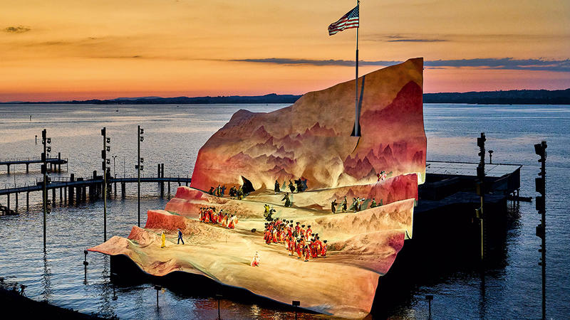 Opernaufführung auf der Bregenzer Seebühne bei Sonnenuntergang vor einer hügeligen Kulisse mit Schauspielern in roten Kostümen