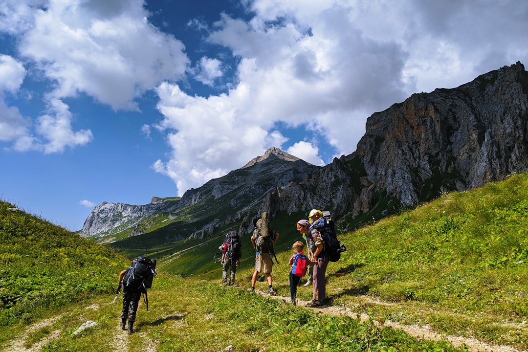 Wandergruppe mit Rucksäcken auf einem Bergpfad vor grasbewachsenen Hängen und schroffen Felsgipfeln unter blauem Wolkenhimmel