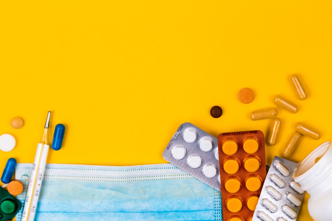 Mundschutz, Thermometer sowie verschiedene Tabletten und Kapseln auf gelbem Hintergrund angeordnet