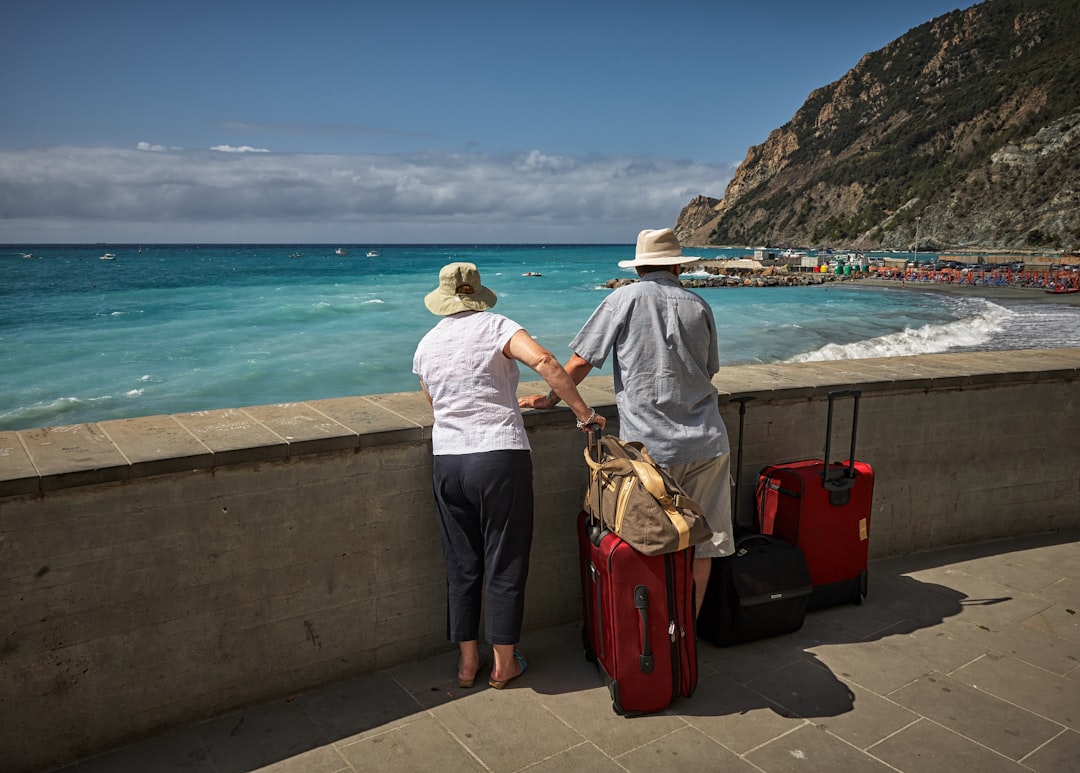 Ein älteres Paar mit Koffern blickt von einer Uferpromenade auf das türkisfarbene Meer und die Küste von Monterosso al Mare